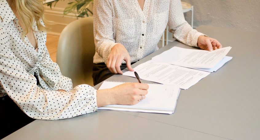 woman signing on white printer paper beside woman about to touch the documents